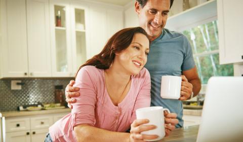 Young adult couple drinking coffee looking at laptop screen in kitchen